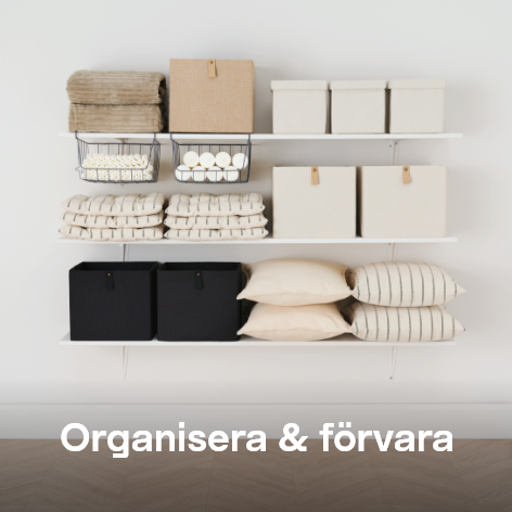 Bild på vita hyllor fyllda med vikta handdukar, förvaringslådor, korgar med ljus och dekorativa kuddar i beige och svart. Text: Organisera & förvara
 