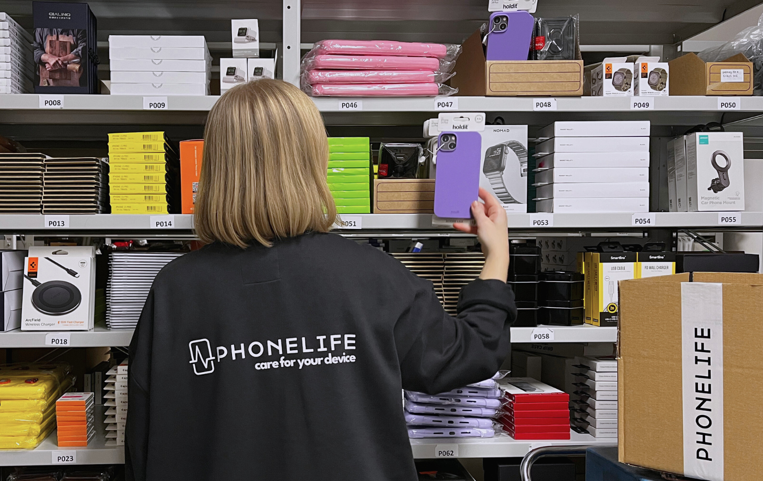 Person med ryggen mot kameran plockar ett lila mobilskal från en lagerhylla fylld med mobiltillbehör. Tröjan har PhoneLife-logotypen på ryggen.