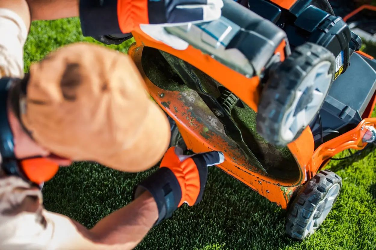 En person i skyddsutrustning inspekterar undersidan och kniven på en orange gräsklippare som är tippad på sidan på en gräsmatta.